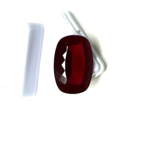 Pode incluir: Uma pedra preciosa vermelho-escura com lapidação almofada e uma superfície facetada que reflete a luz. A pedra tem formato oval e exibe um tom carmesim rico e saturado em todo o seu corpo transparente, com cortes geométricos precisos na superfície.