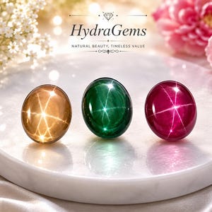 May include: Three star-shaped gemstones in gold, green, and red are displayed on a white marble surface. The gemstones are round and polished, with a star-like pattern visible within each stone. The image includes the text "HydraGems: Natural Beauty, Timeless Value."