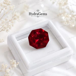 Peut inclure: Une pierre précieuse octogonale à facettes d'un rouge profond, présentée dans une boîte à bijoux carrée blanche. La pierre présente des coupes géométriques nettes qui reflètent la lumière, créant un aspect cramoisi vibrant. Le texte HydraGems est imprimé au-dessus.
