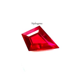 Puede incluir: Una piedra preciosa roja facetada en forma de cometa con apariencia translúcida. La palabra Hydragems está impresa en texto negro sobre la piedra. La gema presenta ángulos geométricos afilados y un color carmesí vibrante y saturado.