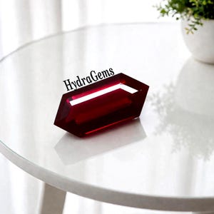 May include: A faceted, deep red gemstone with the text "HydraGems" on top. The gem is displayed on a white surface, reflecting light. The gem's shape is a modified hexagon. A small plant in a white pot is in the background.