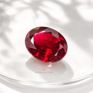 Puede incluir: Una piedra preciosa de color rojo intenso con talla ovalada y múltiples facetas que reflejan la luz. La piedra está situada sobre el texto HYDRAGEMS, impreso en letras pequeñas y oscuras. La gema muestra un color carmesí vibrante y saturado.