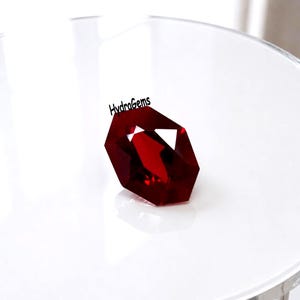 May include: A faceted, deep red gemstone with a geometric shape. The gem is displayed on a white surface, reflecting light. The text "HydraGems" is above the gem.