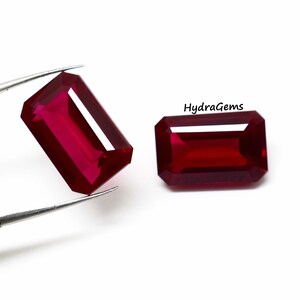 May include: Two radiant, rectangular-cut, deep red gemstones. One is held by tweezers, showcasing its facets. The other gem rests on a white surface. The text "HydraGems" is visible.