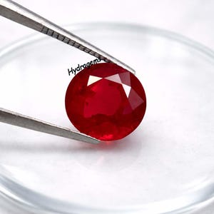 Op de afbeelding: Een gefacetteerde, ronde, dieprode edelsteen wordt vastgehouden door een zilveren pincet. De edelsteen is geslepen om licht te reflecteren. Het woord "Hydragems" staat linksboven. De edelsteen staat tegen een witte achtergrond.