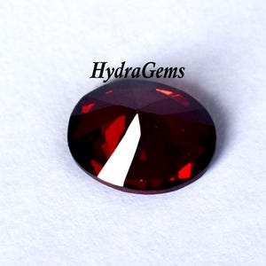 K&ouml;nnte beinhalten: Ein facettierter, oval geformter, tiefroter Edelstein mit einer hellwei&szlig;en Facette. Der Edelstein ist vor einem wei&szlig;en Hintergrund platziert. Der Text "HydraGems" steht in Schwarz &uuml;ber dem Edelstein.