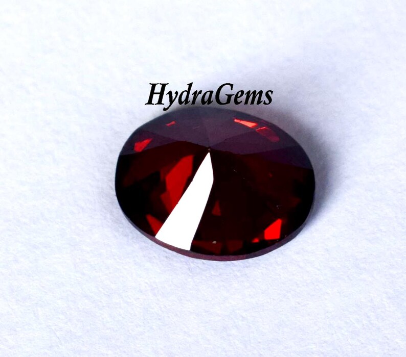 K&ouml;nnte beinhalten: Ein facettierter, oval geformter, tiefroter Edelstein mit einer hellwei&szlig;en Facette. Der Edelstein ist vor einem wei&szlig;en Hintergrund platziert. Der Text "HydraGems" steht in Schwarz &uuml;ber dem Edelstein.
