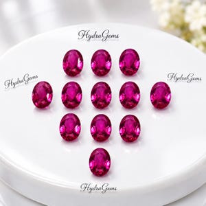 Könnte beinhalten: Eine Ansammlung von elf oval geschliffenen, leuchtend magentafarbenen Edelsteinen, die symmetrisch auf einer glatten weißen Oberfläche angeordnet sind. Jeder facettierte Stein zeigt einen brillanten Glanz und tiefe Farbsättigung. Der Text HydraGems ist sichtbar.