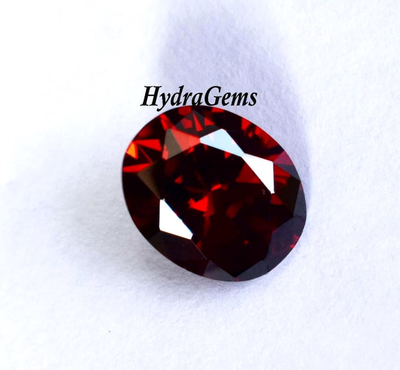K&ouml;nnte beinhalten: Ein facettierter, oval geformter, tiefroter Edelstein. Der Edelstein hat mehrere reflektierende Oberfl&auml;chen, die helle Glanzlichter erzeugen. Der Text "HydraGems" ist in schwarzer, kursiver Schrift &uuml;ber dem Edelstein gedruckt. Der Edelstein befindet sich auf wei&szlig;em Hintergrund.