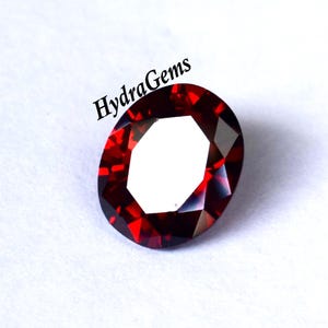 K&ouml;nnte beinhalten: Ein facettierter, oval geformter, tiefroter Edelstein. Der Edelstein hat eine helle, reflektierende Oberfl&auml;che mit einem wei&szlig;en Zentrum. Der Text "HydraGems" ist in Schwarz oben links im Bild gedruckt. Der Edelstein ist vor wei&szlig;em Hintergrund.