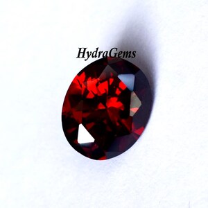 K&ouml;nnte beinhalten: Ein oval geschliffener, tiefroter Edelstein mit Facetten. Der Stein weist eine hohe Klarheit und Brillanz auf, wobei mehrere Facetten das Licht einfangen. Der Text "HydraGems" ist sichtbar.