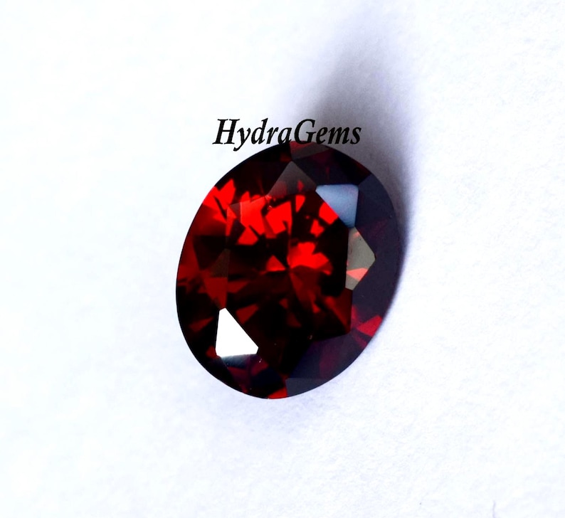 K&ouml;nnte beinhalten: Ein oval geschliffener, tiefroter Edelstein mit Facetten. Der Stein weist eine hohe Klarheit und Brillanz auf, wobei mehrere Facetten das Licht einfangen. Der Text "HydraGems" ist sichtbar.
