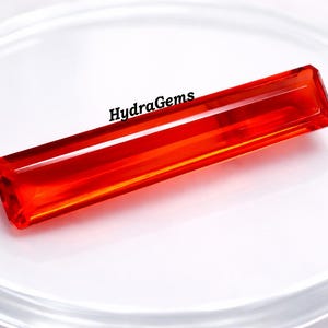 May include: A rectangular, faceted, vibrant orange gemstone. The gem is displayed on a clear, circular surface. The text "HydraGems" is visible on the surface. The gem's color is a rich, saturated orange.