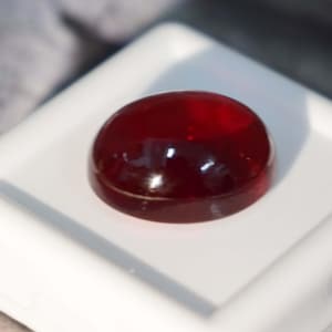May include: A deep red cabochon gemstone, polished and smooth, sits on a white display stand.