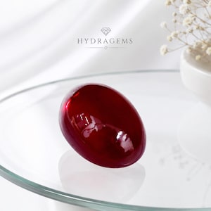 May include: A smooth, polished, deep red oval gemstone with a high-gloss finish. The stone displays a rich, saturated crimson hue with reflective surfaces. Above the stone, the text HYDRAGEMS is printed in a dark font with a diamond icon above it.