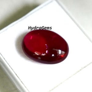 May include: A polished, oval-shaped, deep red gemstone. The gem is set against a white background and reflects light, showcasing its smooth surface. The text "HydraGems" is visible.