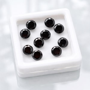 May include: A collection of ten round, black gemstones displayed in a white, square container. The gemstones have a faceted cut, reflecting light. The container is on a reflective surface.