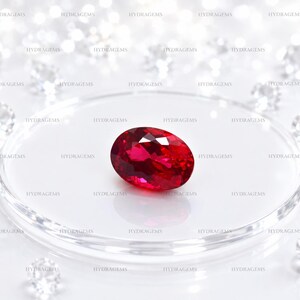 Puede incluir: Una vibrante piedra preciosa de rubí de corte ovalado con una superficie facetada que refleja la luz, exhibida centralmente sobre una plataforma circular de vidrio transparente rodeada de varios diamantes blancos pequeños de corte brillante.