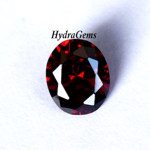 K&ouml;nnte beinhalten: Ein facettierter, oval geformter, tiefroter Edelstein. Der Edelstein hat eine hohe Klarheit mit mehreren reflektierenden Oberfl&auml;chen. Der Text "HydraGems" ist sichtbar.