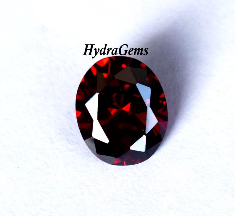K&ouml;nnte beinhalten: Ein facettierter, oval geformter, tiefroter Edelstein. Der Edelstein hat eine hohe Klarheit mit mehreren reflektierenden Oberfl&auml;chen. Der Text "HydraGems" ist sichtbar.
