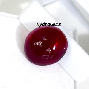 Op de afbeelding: Een ronde, gepolijste, dieprode edelsteen met een glanzend oppervlak. De edelsteen is geplaatst tegen een witte achtergrond. De tekst "HydraGems" is zichtbaar in de rechterbovenhoek.