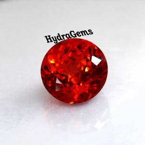 Puede incluir: Una gema roja vibrante, facetada y redonda. La gema tiene un corte brillante, que refleja la luz con múltiples facetas. El texto "HydraGems" está impreso encima de la gema. La gema está sobre un fondo blanco.