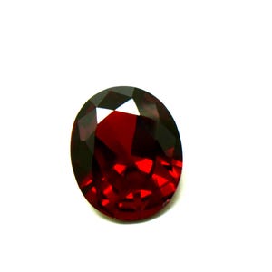 May include: A deep red, oval-shaped gemstone with a faceted cut. The stone features a brilliant, transparent surface with multiple geometric facets that reflect light, creating a vibrant, saturated crimson appearance throughout the gem.