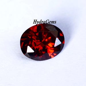 K&ouml;nnte beinhalten: Ein facettierter, ovalf&ouml;rmiger, tiefroter Edelstein. Der Edelstein hat einen Brillantschliff, der das Licht mit hellen Blitzen reflektiert. Der Text "HydraGems" ist oben sichtbar. Der Edelstein befindet sich vor einem wei&szlig;en Hintergrund.