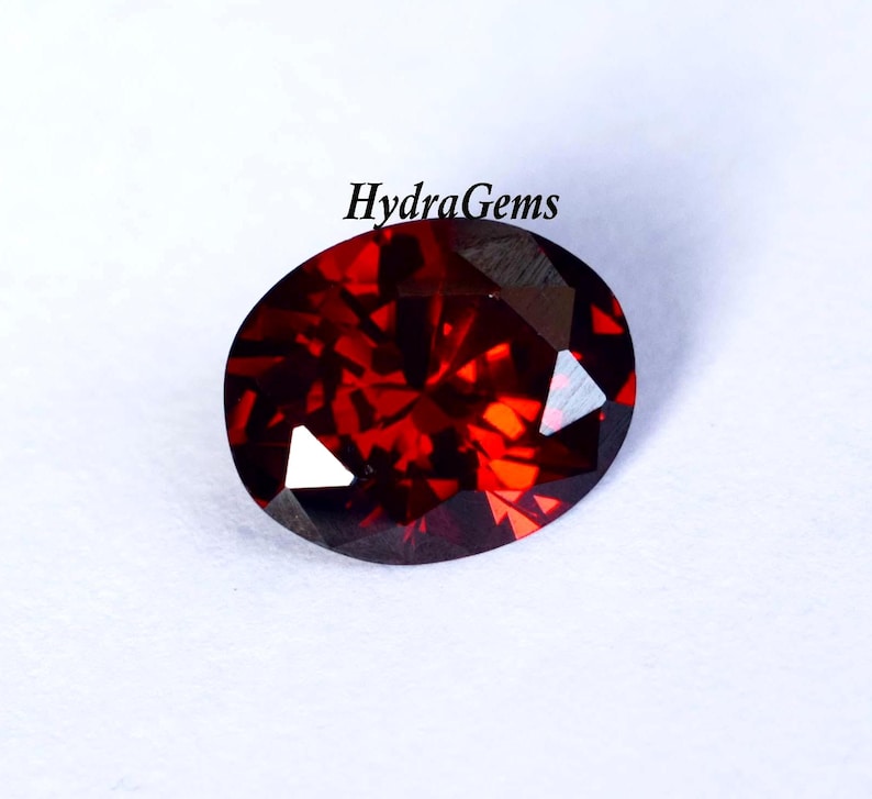 K&ouml;nnte beinhalten: Ein facettierter, ovalf&ouml;rmiger, tiefroter Edelstein. Der Edelstein hat einen Brillantschliff, der das Licht mit hellen Blitzen reflektiert. Der Text "HydraGems" ist oben sichtbar. Der Edelstein befindet sich vor einem wei&szlig;en Hintergrund.