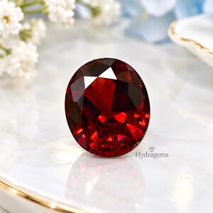 Könnte beinhalten: Ein tiefroter, oval geschliffener Edelstein mit einer facettierten Oberfläche, die das Licht reflektiert. Der Stein zeigt einen satten, kräftigen karmesinroten Farbton. Der Text Hydragems ist in kleiner schwarzer Schrift unten rechts gedruckt.