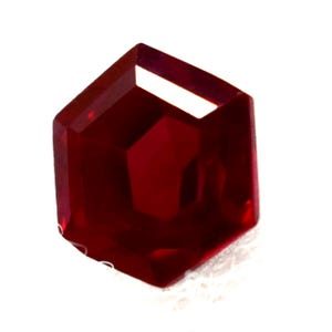 Puede incluir: Una piedra preciosa facetada de color rojo intenso con forma hexagonal. La piedra presenta múltiples superficies planas y pulidas que reflejan la luz, creando una apariencia translúcida con varios tonos carmesí en toda la estructura geométrica.