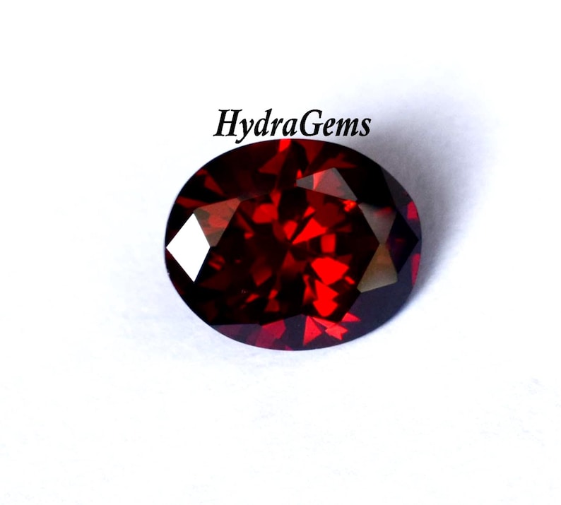 K&ouml;nnte beinhalten: Ein facettierter, oval geformter, tiefroter Edelstein. Der Edelstein hat einen Brillantschliff, der das Licht mit mehreren Facetten reflektiert. Der Text "HydraGems" ist im Bild enthalten.