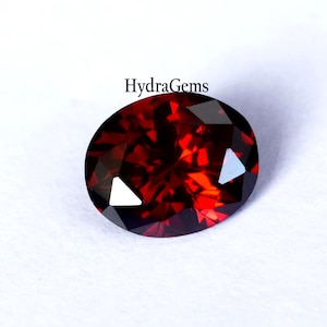K&ouml;nnte beinhalten: Ein facettierter, oval geformter, tiefroter Edelstein. Der Edelstein hat einen Brillantschliff mit mehreren Facetten, die das Licht reflektieren. Der Text "HydraGems" ist im Bild enthalten. Der Edelstein ist vor einem wei&szlig;en Hintergrund platziert.