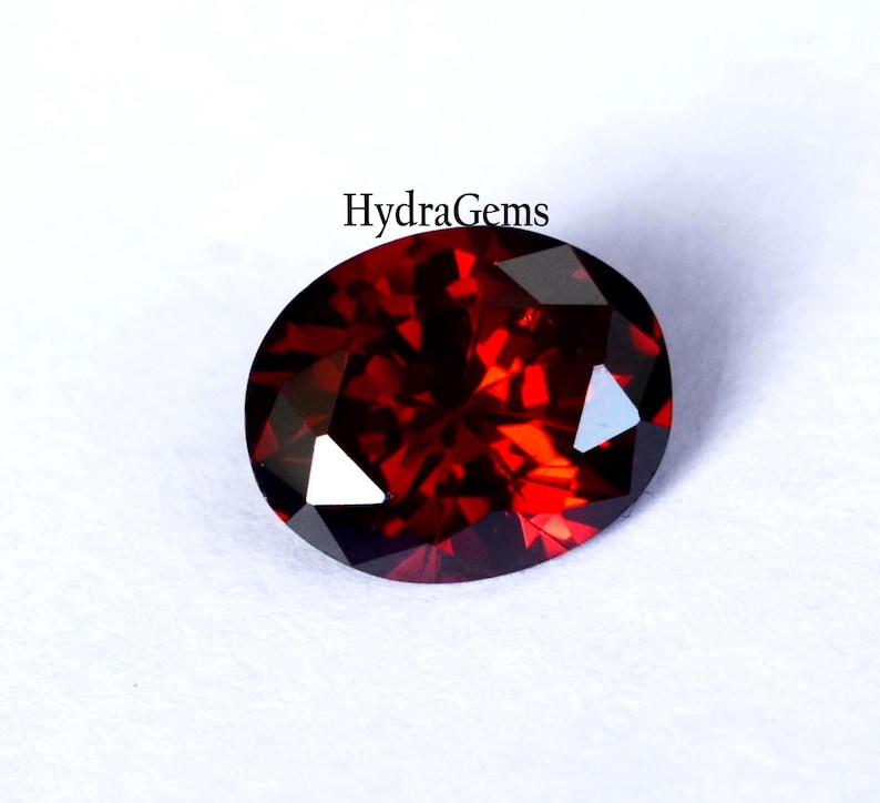 K&ouml;nnte beinhalten: Ein facettierter, oval geformter, tiefroter Edelstein. Der Edelstein hat einen Brillantschliff mit mehreren Facetten, die das Licht reflektieren. Der Text "HydraGems" ist im Bild enthalten. Der Edelstein ist vor einem wei&szlig;en Hintergrund platziert.