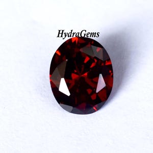 K&ouml;nnte beinhalten: Ein oval geschliffener, tiefroter Edelstein. Der Edelstein hat einen Brillantschliff und reflektiert das Licht. Der Text "HydraGems" befindet sich oben rechts. Der Edelstein ist vor wei&szlig;em Hintergrund.