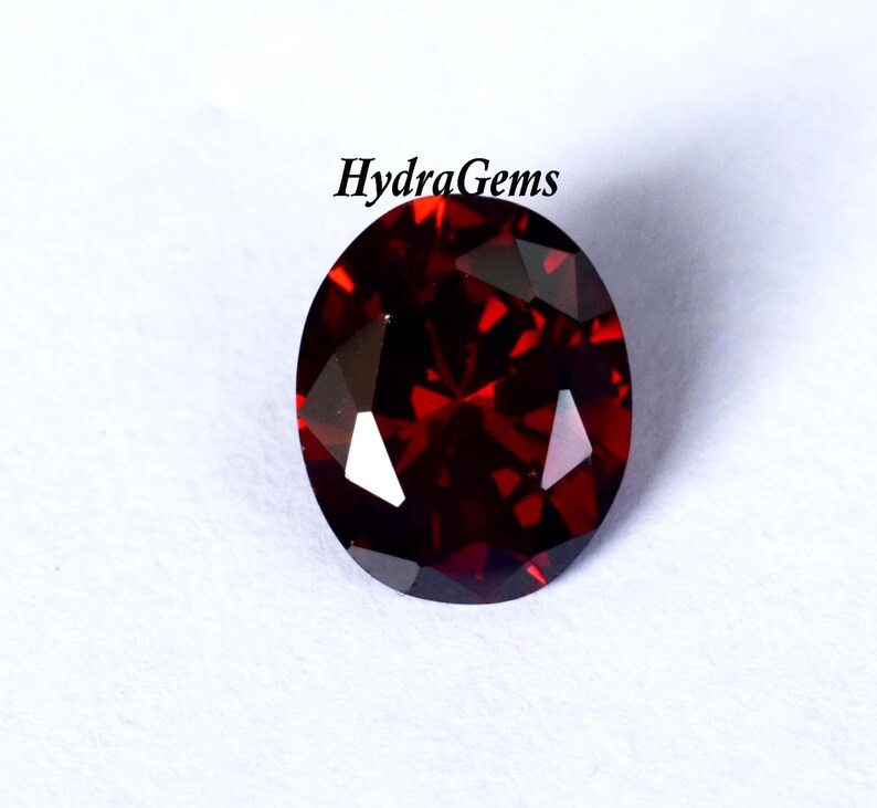 K&ouml;nnte beinhalten: Ein oval geschliffener, tiefroter Edelstein. Der Edelstein hat einen Brillantschliff und reflektiert das Licht. Der Text "HydraGems" befindet sich oben rechts. Der Edelstein ist vor wei&szlig;em Hintergrund.