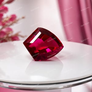 Puede incluir: Una gema de rubí tallada en forma cuadrada, de color rojo intenso. La gema se exhibe sobre una superficie blanca y reflectante, con un fondo rosa suave. Las facetas de la gema capturan la luz, creando un brillo brillante.