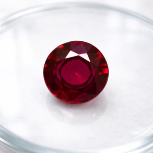 Puede incluir: Una gema de rubí redonda y facetada. La gema es de color rojo intenso con una superficie brillante y reflectante. El rubí se encuentra sobre un fondo blanco, resaltando su color y claridad. La piedra preciosa es un símbolo de amor y pasión.