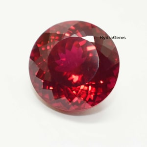 Puede incluir: Una gema facetada, redonda, de color rojo intenso. La gema tiene un corte brillante con muchas superficies reflectantes, creando un efecto brillante. El texto "HydraGems" es visible. Adecuada para la fabricación de joyas.