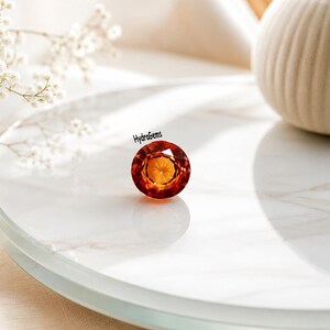 May include: A round, faceted gemstone in shades of orange and yellow. The gem is displayed on a white surface, with the text "HydraGems" visible. This gem could be used in jewellery.