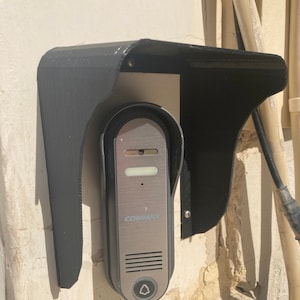 May include: A silver door intercom with a black plastic weather shield. The intercom has a button with a bell icon and the word "COMMAX" printed on it.