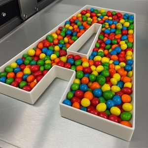 May include: A white plastic letter A-shaped container filled with a variety of small, round, multi-coloured sweets. The sweets are red, orange, yellow, green, and blue, creating a vibrant display within the structured, hollow letter frame.