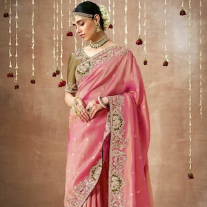 May include: A pink silk saree with intricate gold embroidery and a green and gold border. The saree is draped on a woman wearing a green blouse.