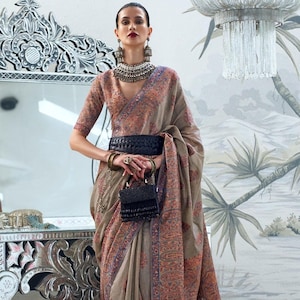 May include: A woman wearing a beige and blue patterned saree with a black belt and a black beaded purse. She is wearing silver jewelry and has her hair pulled back.