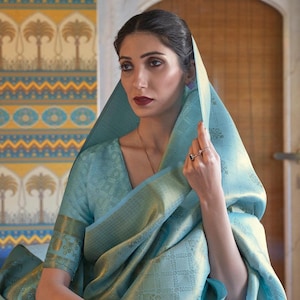 May include: A woman wearing a light blue and gold saree with a gold trim. She is holding a small, light brown clutch with a gold elephant design.