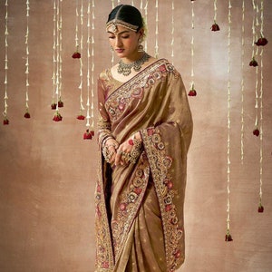 May include: A gold and maroon saree with intricate embroidery and embellishments. The saree has a floral design and is draped on a model.