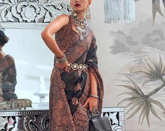 May include: A woman wearing a black and brown patterned saree with a silver belt and a black handbag. She is wearing silver jewelry and has her hair pulled back.