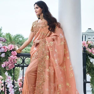 May include: A peach-coloured saree with gold embroidery and floral patterns. The saree has a beaded border and a matching blouse.