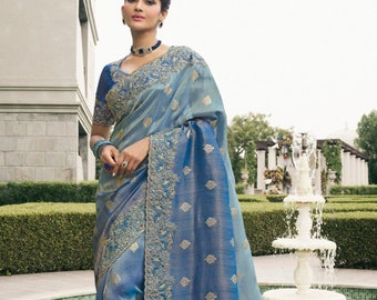 May include: A blue and silver saree with intricate embroidery and a delicate border. The saree is draped on a woman standing in front of a fountain.