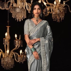 Puede incluir: Un sari gris plateado con bordados intrincados y un borde de cuentas. El sari está drapeado sobre una modelo y presenta una blusa gris plateado con un diseño de cuentas similar.